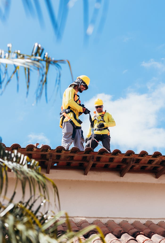 Two construction workers in safety gear working on a tiled roof under a clear blue sky.